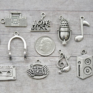 May include: A collection of silver-toned metal charms with music-themed designs, including a cassette tape, a boombox, a microphone, a treble clef, a music note, headphones, and a speaker. The charms are arranged on a wooden surface.