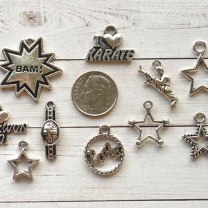 May include: A collection of silver-toned charms featuring various designs, including stars, a karate charm with the text "I love karate", a charm with the text "BAM!" and a charm with the text "Tae-kwon Do!"