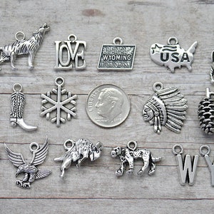 May include: A collection of 18 silver charm pendants featuring various designs, including a cowboy boot, a snowflake, a pine cone, a wolf, a buffalo, a deer, a cowboy hat, a gun, an eagle, a bear, a mountain lion, a Native American headdress, the words "LOVE", "USA", and "WY", and a state of Wyoming outline.
