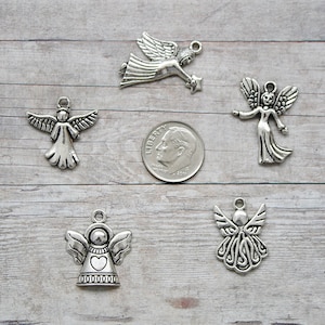 K&ouml;nnte beinhalten: Sechs silberne Engelscharms mit verschiedenen Designs, darunter ein Herz, Fl&uuml;gel und ein Stern. Die Charms sind auf einer hellbraunen Holzoberfl&auml;che mit einer deutschen Zehn-Cent-M&uuml;nze zur Gr&ouml;&szlig;enbestimmung angeordnet.