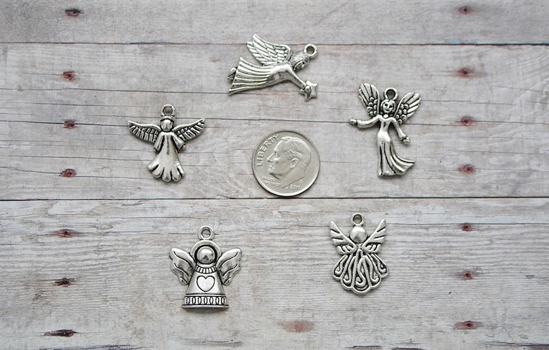 K&ouml;nnte beinhalten: Sechs silberne Engelscharms mit verschiedenen Designs, darunter ein Herz, Fl&uuml;gel und ein Stern. Die Charms sind auf einer hellbraunen Holzoberfl&auml;che mit einer deutschen Zehn-Cent-M&uuml;nze zur Gr&ouml;&szlig;enbestimmung angeordnet.