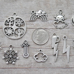 May include: A collection of silver-toned weather-themed charms, including an umbrella, sun, snowflake, rainbow, and lightning bolt. The charms are displayed on a wooden surface, with a dime for size comparison. Ideal for jewelry making.