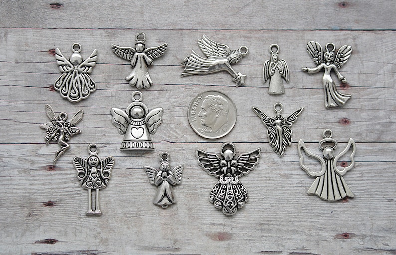 K&ouml;nnte beinhalten: Eine Sammlung von 13 silbernen Engelsanh&auml;ngern, jeder mit einem einzigartigen Design. Die Anh&auml;nger sind auf einer Holzoberfl&auml;che angeordnet, mit einer 10-Cent-M&uuml;nze als Gr&ouml;&szlig;envergleich.