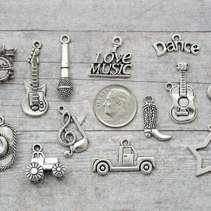 May include: A collection of silver charm pendants featuring musical instruments, a cowboy hat, a truck, a star, a boot, and a treble clef. The charms are arranged on a wooden surface. The charms are all silver in color and have a rustic, vintage look.