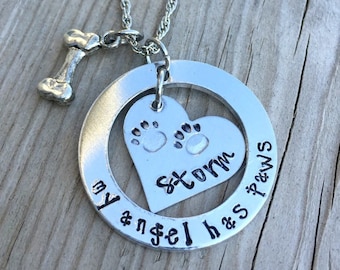 以下が含まれることがあります： 「my angel has paws」と書かれた丸いペンダントと、中に「Storm」という名前と肉球の跡が入ったハート型の切り抜きが付いたシルバーネックレス。ペンダントの上部から小さな骨のチャームがぶら下がっている。