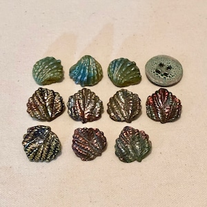 May include: Assortment of 11 iridescent glass buttons in various shades of green and brown. The buttons are shaped like seashells and leaves.