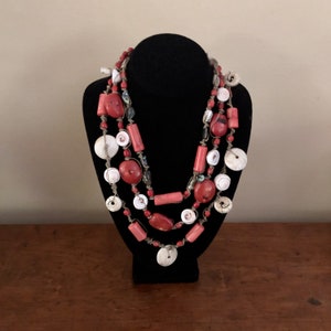 Puede incluir: Un collar de múltiples hilos con cuentas de coral rojo y conchas blancas. El collar está hecho con materiales naturales y tiene un estilo rústico y bohemio.