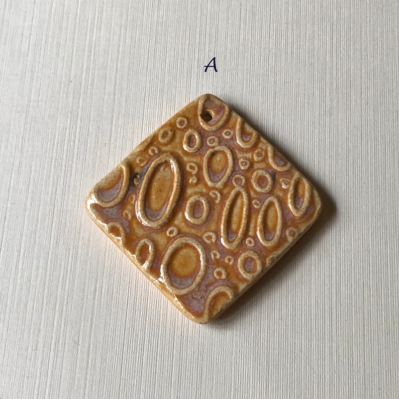 May include: A brown ceramic tile with a raised pattern of circles and ovals. The tile is in the shape of a diamond.