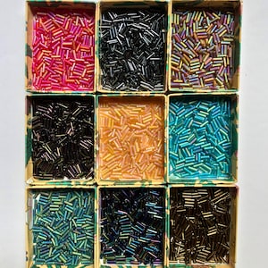 以下が含まれることがあります： カラフルな円筒形のビーズが詰められた9つの小さな段ボール箱。ビーズはピンク、黒、金、虹色の青と緑など、さまざまな色があります。箱の縁には花柄があります。