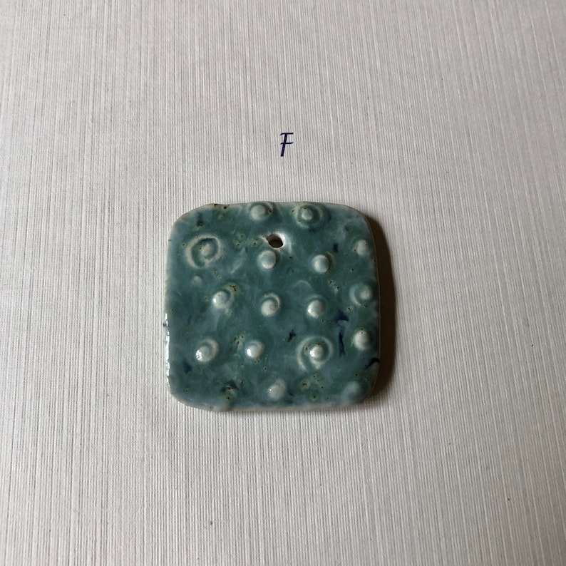 May include: A square ceramic tile with a textured surface. The tile is a light blue color with white dots and swirls. The tile has a hole in the top center.