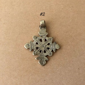 May include: A silver-toned metal pendant in the shape of a cross with intricate details. The cross has a circular design in the center and four arms that extend outwards. The pendant has a loop at the top for attaching to a chain.