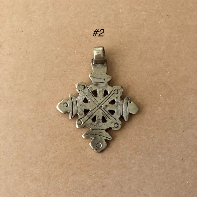 May include: A silver-toned metal pendant in the shape of a cross with intricate details. The cross has a circular design in the center and four arms that extend outwards. The pendant has a loop at the top for attaching to a chain.