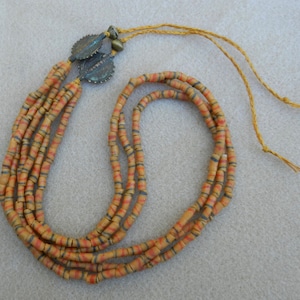 Puede incluir: Un collar de múltiples hilos hecho de pequeñas cuentas cilíndricas con rayas naranjas y negras alternas. El collar tiene un borla hecho de un cordón fino marrón con dos pequeños abalorios metálicos redondos en la parte superior.
