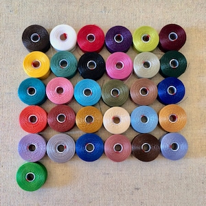 以下が含まれることがあります： 整然と並んだ、カラフルなワックス糸の糸巻きのコレクション。糸巻きは、茶色、白、赤、紫、黄色、緑、黒、ピンク、青、金など、さまざまな色を表示しています。糸は各糸巻きにしっかりと巻かれています。