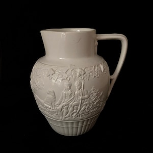 May include: White ceramic pitcher with an embossed design of three figures and a dog in a wooded setting. The pitcher has a rounded body and a handle.