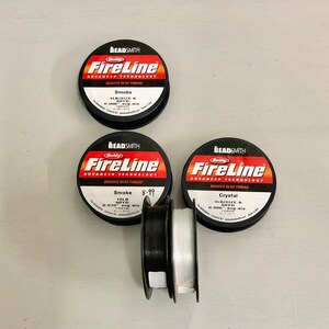 May include: Three spools of Berkley FireLine braided bead thread. The spools are black with red and white text. The thread colors are smoke, smoke, and crystal. The spools are on a white surface.