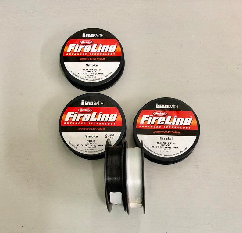 May include: Three spools of Berkley FireLine braided bead thread. The spools are black with red and white text. The thread colors are smoke, smoke, and crystal. The spools are on a white surface.