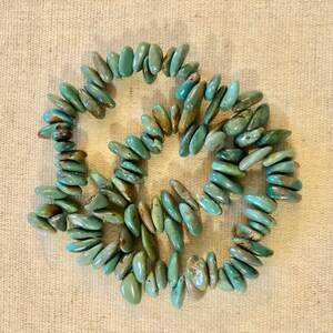 May include: A necklace composed of turquoise-coloured, oblong beads. The beads are a mix of light and dark teal, with some showing brown and tan hues. The necklace is laid out on a neutral-coloured, textured surface.