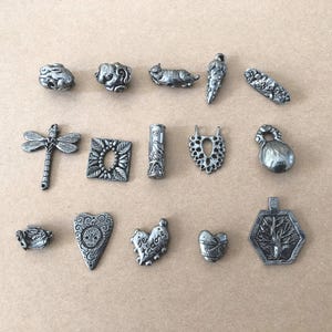 May include: A collection of small, silver-colored metal charms and pendants. The charms include a dragonfly, a cat, a heart with the words "Je t'adore", and other decorative shapes. The items are arranged on a light brown surface.