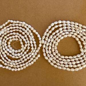 May include: Two strands of white oval-shaped freshwater pearls. The pearls are strung on a white thread and are arranged in a circular pattern.