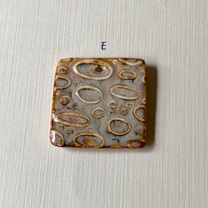 May include: Square ceramic tile with a textured surface. The tile has a light gray background with brown and white oval shapes. The tile has a hole in the top for hanging.