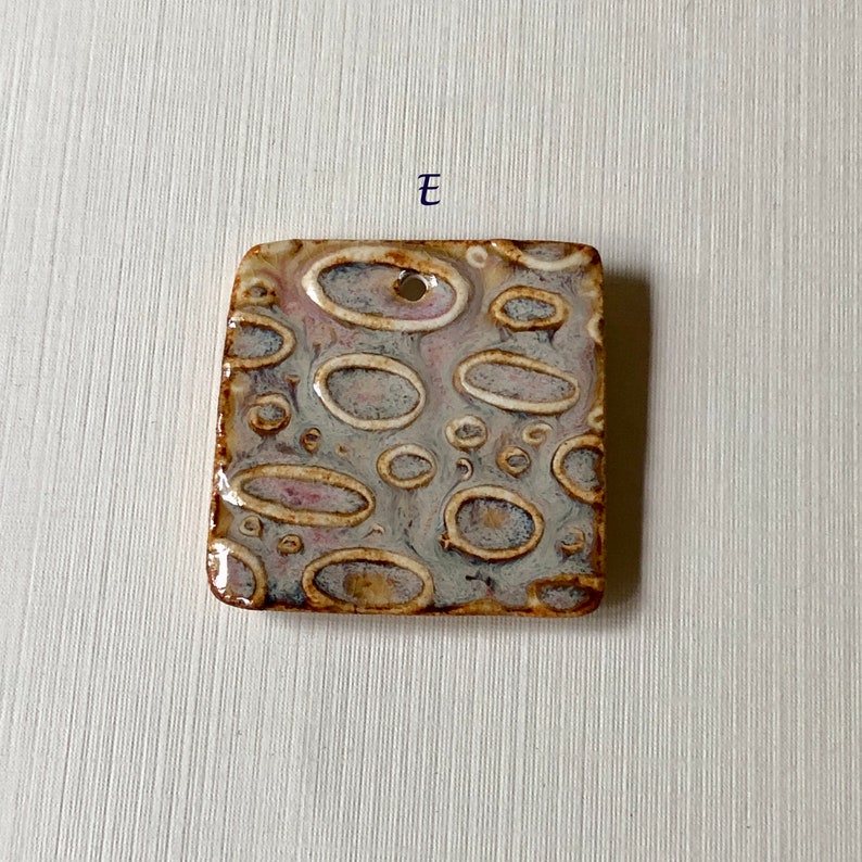 May include: Square ceramic tile with a textured surface. The tile has a light gray background with brown and white oval shapes. The tile has a hole in the top for hanging.