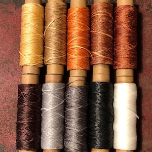 May include: Ten spools of waxed thread in various colors, including gold, brown, orange, and black. The spools are arranged in two rows, with the thread wound around cardboard tubes. The thread is likely used for leathercraft or other crafting projects.