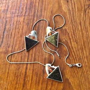 Puede incluir: Tres collares de plata con colgantes triangulares. Cada colgante tiene una superficie oscura y texturizada y un acento plateado con una pequeña gema naranja. Los collares se exhiben sobre una superficie de madera.