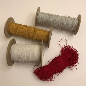 May include: Four spools of thread, one each in gold, white, red, and silver. The spools are on a white surface.