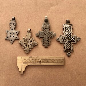 May include: Four silver pendants, including a star of David and three crosses, are displayed on a brown surface. A ruler is included for scale.