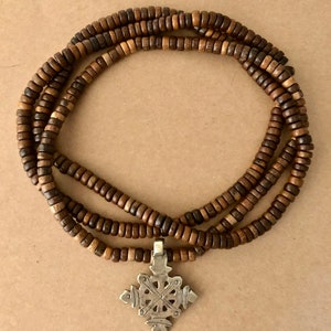 May include: A brown wooden beaded necklace with a silver pendant in the shape of a cross. The pendant has a detailed design with intricate lines and curves.