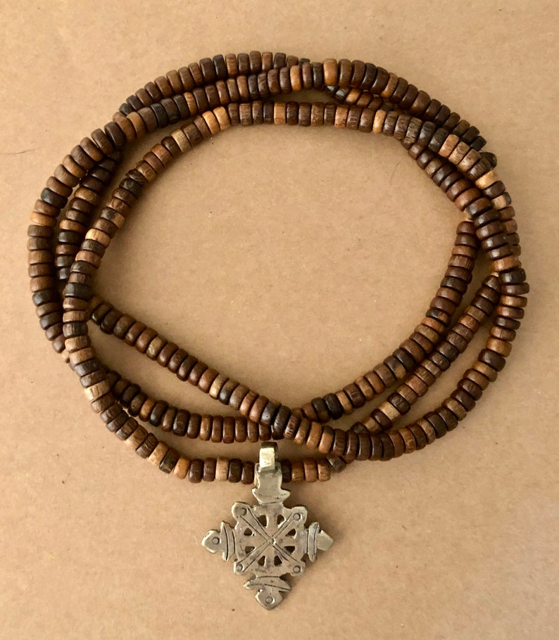 May include: A brown wooden beaded necklace with a silver pendant in the shape of a cross. The pendant has a detailed design with intricate lines and curves.