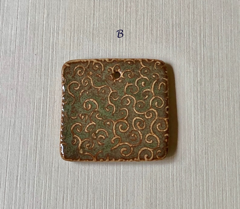 May include: A square ceramic tile with a green and brown glaze. The tile has a decorative pattern of swirls and is edged with a brown border. The tile has a small hole in the center.