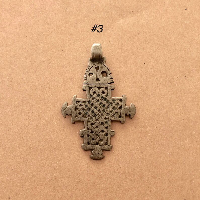 May include: A silver-toned cross pendant with intricate designs. The cross is made of metal and has a loop at the top for hanging.