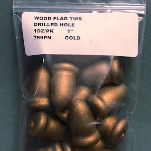 以下が含まれることがあります： 金色の木製フラッグチップが入った透明なビニール袋。「WOOD FLAG TIPS DRILLED HOLE 1DZ/PK 2.54cm GOLD 759PN」と書かれたラベル。チップは円筒形で、上部が丸くなっています。