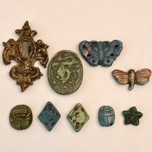Könnte beinhalten: Eine Sammlung von neun dekorativen Keramikknöpfen mit verschiedenen Designs, darunter ein Kreuz, ein Schmetterling, eine Libelle, eine Rose, ein Stern und ein Skarabäus. Die Knöpfe sind in Grün-, Blau- und Brauntönen bemalt.