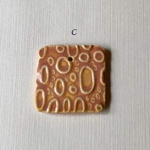 May include: A square ceramic tile with a textured surface. The tile is decorated with a pattern of circles and ovals in shades of orange and yellow.