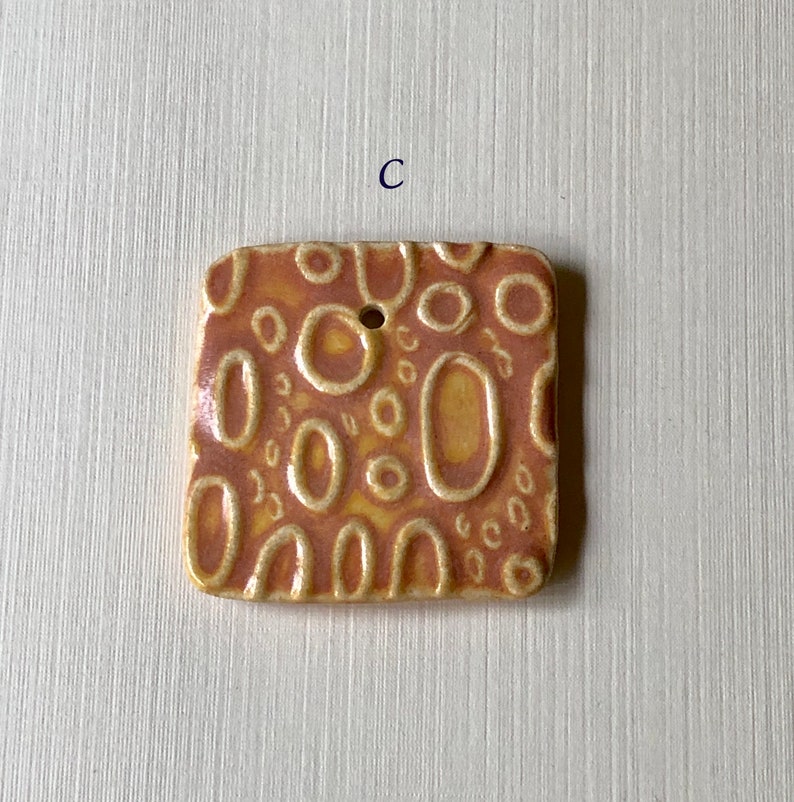 May include: A square ceramic tile with a textured surface. The tile is decorated with a pattern of circles and ovals in shades of orange and yellow.