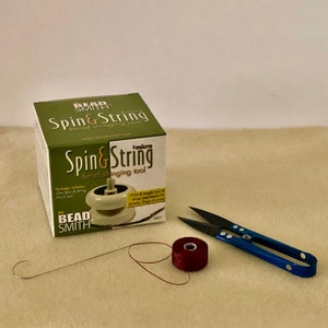 Puede incluir: Un kit para ensartar cuentas con una caja verde y blanca con la etiqueta "Spin & String". El kit incluye una microherramienta, un carrete de hilo rojo, unas tijeras con mango azul y una aguja de alambre.