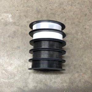 May include: Five spools of thread, four black and one white, stacked on top of each other. The spools are all the same size and have a black plastic base.
