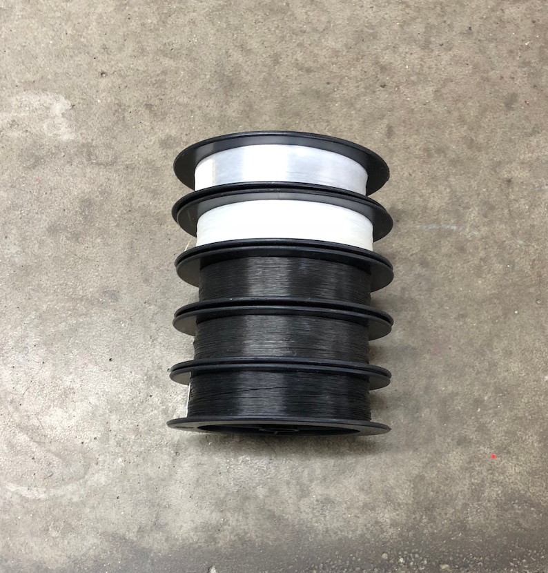 May include: Five spools of thread, four black and one white, stacked on top of each other. The spools are all the same size and have a black plastic base.