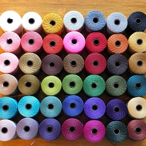 May include: A collection of 49 spools of thread in various colors, including white, pink, red, brown, green, blue, and gray. The spools are arranged in a grid pattern on a wooden surface.