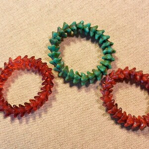 May include: Three beaded bracelets. Two are red, and one is green. Each bracelet is made of small, pointed beads strung together in a circular shape. The bracelets are displayed on a neutral-colored fabric.