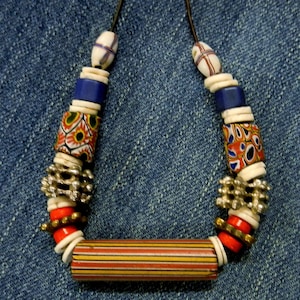 May include: A handcrafted necklace featuring a variety of colorful beads. The necklace includes cylindrical beads with red, yellow, and orange stripes, along with patterned and solid blue beads. The beads are strung on a dark cord.
