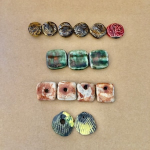 May include: Assortment of ceramic beads in various colors and patterns, including green, brown, yellow, and red. The beads are arranged in three rows, with the top row featuring round beads, the middle row featuring square beads, and the bottom row featuring two shell-shaped beads.