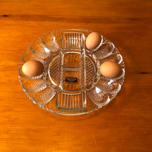 May include: A clear glass devilled egg serving dish with a central compartment and egg-shaped indentations. The dish holds three brown eggs. The dish has a Heritage label.