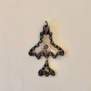 Puede incluir: Un colgante de plata con forma triangular y un colgante más pequeño que cuelga de la parte inferior. El colgante está decorado con pequeñas cuentas redondas.