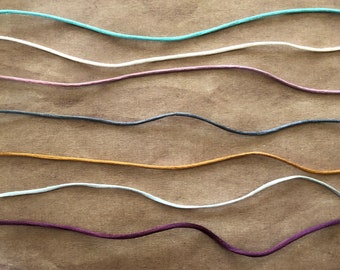 May include: A collection of eight different colored cords, arranged in a wavy pattern on a brown surface. The cords are all the same thickness and are made of a soft, pliable material.
