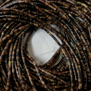 Puede incluir: Una hebra de cuentas de concha marrón y blanca. Las cuentas son pequeñas y cilíndricas, y están enhebradas para formar un collar largo.