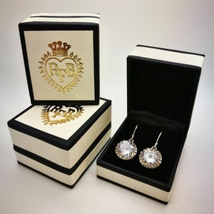 May include: Earrings with large, round, clear gemstones are displayed in a black velvet-lined box. The box is cream-colored with black trim and a gold logo. Two additional cream-colored boxes with black trim and gold logos are also visible.